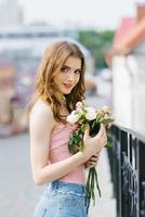 una hermosa joven de pelo rubio con ropa de verano se para y sostiene delicadas rosas blancas y rosadas en sus manos después de una cita foto