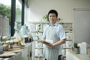 Portrait of an Asian male shopkeeper smiling and looking at camera, arranges natural products at refill store, zero-waste grocery, and plastic-free, eco environment-friendly, sustainable lifestyles. photo
