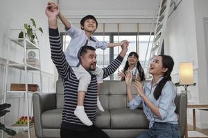 familia asiática tailandesa unida, papá juega y bromea con los niños y la mamá cargando y sosteniendo a su hijo sobre los hombros en la sala de estar de la casa, tiempos de ocio felices, fin de semana encantador, estilo de vida doméstico de bienestar. foto