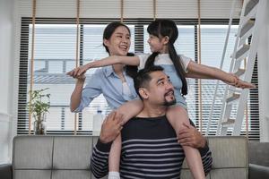 Asian Thai family together, dad plays and teases with daughter and mum by carrying and holding girl on shoulders in home living room, happy leisure times, lovely weekend, wellbeing domestic lifestyle. photo