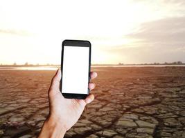 mano de hombre sosteniendo un smartphone negro con pantalla en blanco detrás de tierra de suelo agrietado con fondo borroso. espacio para texto o diseño foto