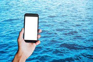 Young man hand holding black smartphone with blank screen behind of view of blue ocean waves background. Space for text or design photo