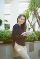 retrato de una estudiante sonriente leyendo un libro mientras está de pie al aire libre en la terraza del café del campus en un día soleado. concepto de educación, estilo de vida y personas. foto