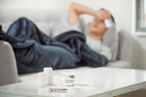 sick wasted man lying in sofa suffering cold and winter flu virus having medicine tablets in health care concept looking temperature on thermometer photo