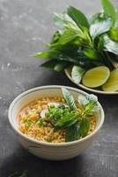 Instant noodles in bowl with fresh herbs, garnish of cilantro and Asian basil, lemon, lime on dark stone background photo