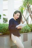 Portrait of smiling female student reading book while standing outdoor on terrace of campus cafe in sunny day. Education, lifestyle and people concept. photo