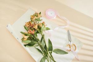 top view of beautiful orange flowers, scissors, ribbon and craft paper on paper surface photo