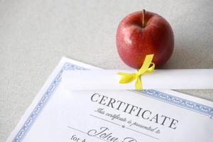 A certificate of achievement lies on table with small scroll and red apple. Education documents photo