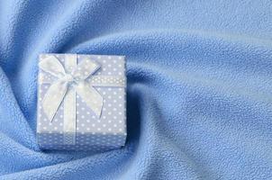 A small gift box in blue with a small bow lies on a blanket of soft and furry light blue fleece fabric with a lot of relief folds. Packing for a gift to your lovely girlfriend photo