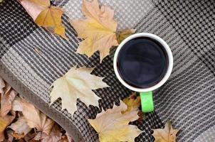 hojas de otoño y una taza de café caliente y humeante se encuentran en cuadros escoceses al aire libre foto