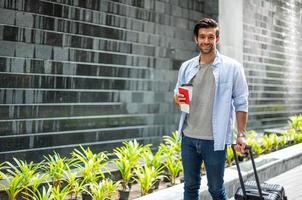 Young caucasian man holding the coffee cup and pulling the suitcase waiting his friend for travel together. photo