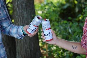 SUMY, UKRAINE - AUGUST 01, 2021 Young man raise Budweiser Bud beer can with male friend on blurred river with kayak and trees. Budweiser is one of the most popular beer brands in the USA photo