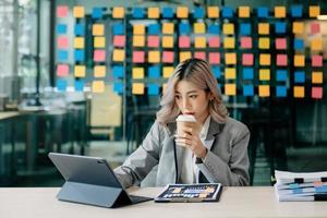 confiada hermosa mujer de negocios asiática escribiendo computadora portátil y tableta digital mientras toma café en la oficina foto