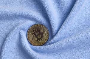 The golden bitcoin lies on a blanket made of soft and fluffy light blue fleece fabric with a large number of relief folds. The shape of the folds resembles a fan from a video card cooler photo