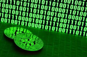 A pair of bitcoins lies on a cardboard surface on the background of a monitor depicting a binary code of bright green zeros and one units on a black background. Low key lighting photo