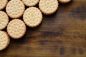 A round sandwich cookie with coconut filling lies in large quantities on a brown wooden surface. Photo of edible treats on a wooden background with copy space