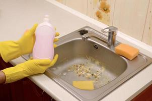 el limpiador muestra una botella de detergente líquido en el fregadero de la cocina sucia con partículas de comida antes de la limpieza foto