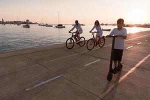 familia feliz disfrutando juntos de una hermosa mañana junto al mar, padres montando en bicicleta y su hijo montando una scooter eléctrica. enfoque selectivo foto