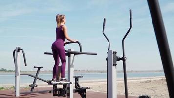 mulher atlética magra em um exercício de agasalho esportivo roxo em uma bicicleta estacionária no campo de treino em um parque da cidade. fitness ao ar livre. câmera lenta video