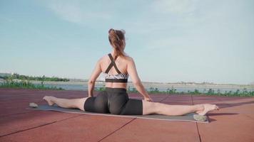 jeune femme athlétique assise en grand écart et faisant du yoga sur fond de lac. belle fille fait de l'exercice à l'extérieur. exercices d'étirement et d'assouplissement. ralenti