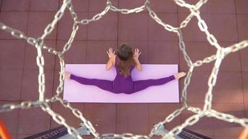 Young athletic woman in a sports purple tracksuit does exercise for stretching while sitting in a split. Fitness outdoors. Slow motion. Top view through a basketball hoop video