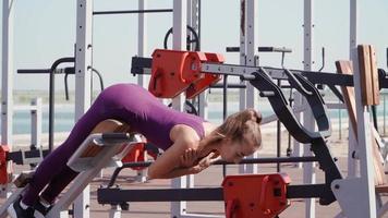 jovem mulher atlética em um agasalho esportivo roxo faz exercícios de hiperextensão lombar na área de treino em um parque da cidade. fitness ao ar livre. câmera lenta video
