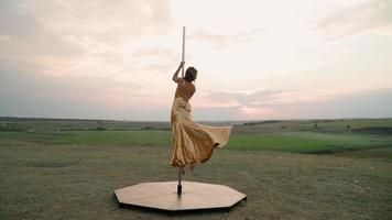 hermosa mujer atlética con un vestido dorado realiza trucos en el poste contra el fondo de la puesta de sol en el campo. belleza y cuidado del cuerpo. deportes femeninos y fitness. camara lenta video