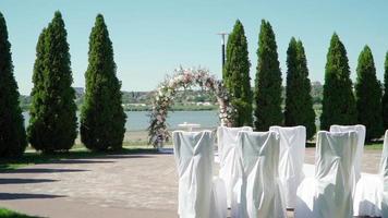 Place for bridal ceremony in white color against the background of the river. Wedding arch with bouquets of pastel roses. Slow motion video