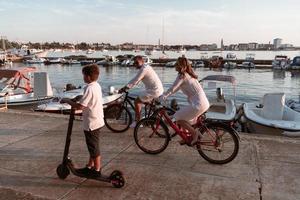 familia feliz disfrutando juntos de una hermosa mañana junto al mar, padres montando en bicicleta y su hijo montando una scooter eléctrica. enfoque selectivo foto