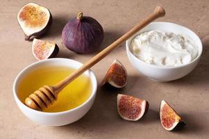 Cut fresh figs, honey in white bowl with wooden spoon and cream cheese on table. photo