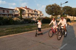 familia feliz disfrutando juntos de una hermosa mañana junto al mar, padres montando en bicicleta y su hijo montando una scooter eléctrica. enfoque selectivo foto