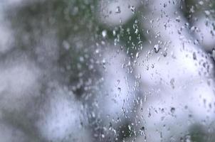 A photo of rain drops on the window glass with a blurred view of the blossoming green trees. Abstract image showing cloudy and rainy weather conditions