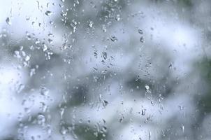 A photo of rain drops on the window glass with a blurred view of the blossoming green trees. Abstract image showing cloudy and rainy weather conditions