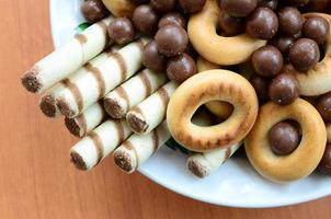 túbulos crujientes, bolas de fusión de chocolate y bagels yacen en un plato blanco sobre una mesa de madera. mezcla de varios dulces foto