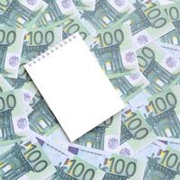 White notebook with clean pages lying on a set of green monetary denominations of 100 euros. A lot of money forms an infinite heap photo