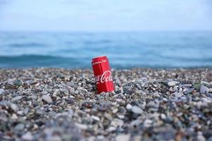 antalya, turquía - 18 de mayo de 2021 la lata roja original de coca cola se encuentra sobre pequeñas piedras redondas cerca de la orilla del mar. coca-cola en playa turca foto