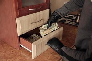 ladrón con traje negro y guantes se ve en un estante abierto en la cocina. el ladrón saca los billetes de dólar estadounidense de un estante foto