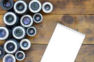 Several photographic lenses and white notebook lie on a brown wooden background. Space for text photo