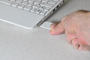 una mano masculina inserta una tarjeta sd compacta blanca en la entrada correspondiente en el lateral de la netbook blanca. el hombre utiliza tecnologías modernas para almacenar memoria y datos digitales foto