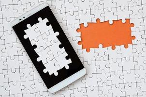 A modern big smartphone with several puzzle elements on the touch screen lies on a white jigsaw puzzle in an assembled state with missing elements photo
