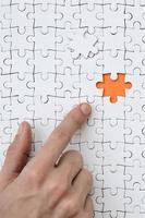 The texture of a white jigsaw puzzle in the assembled state with one missing element, forming an orange space, pointed to by the finger of the male hand photo