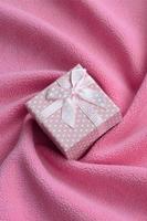 una pequeña caja de regalo de color rosa con un pequeño lazo se encuentra sobre una manta de tela suave y peluda de color rosa claro con muchos pliegues en relieve. embalaje para un regalo a su encantadora novia foto