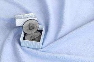The silver bitcoin lies in a small blue gift box with a small bow on a blanket made of soft and fluffy light blue fleece fabric with a large number of relief folds photo