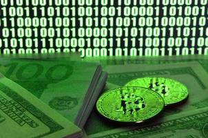 Two bitcoins lies on a pile of dollar bills on the background of a monitor depicting a binary code of bright green zeros and one units on a black background. Low key lighting photo