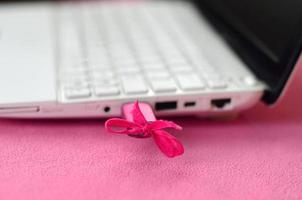 A brilliant pink USB flash drive with a pink bow is connected to a white laptop, which lies on a blanket of soft and fluffy light pink fleece fabric. Classic female design for a memory card photo