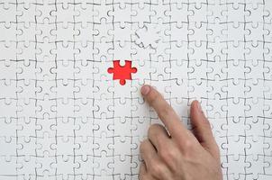 The texture of a white jigsaw puzzle in the assembled state with one missing element, forming a red space, pointed to by the finger of the male hand photo