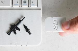 una mano inserta una tarjeta sd compacta en la ranura de un netbook, en la que se encuentran un cuchillo y una pistola en miniatura. el concepto de guerra de información, actividad periodística, difusión de información provocativa foto