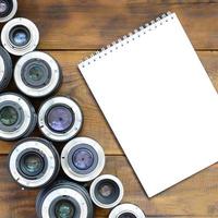 Several photographic lenses and white notebook lie on a brown wooden background. Space for text photo