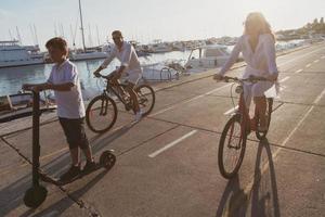 familia feliz disfrutando juntos de una hermosa mañana junto al mar, padres montando en bicicleta y su hijo montando una scooter eléctrica. enfoque selectivo foto