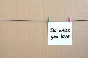 Do what you love. Note is written on a white sticker that hangs with a clothespin on a rope on a background of brown cardboard photo